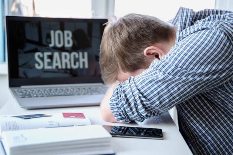 Man lying face down on table next to laptop, feeling no confidence