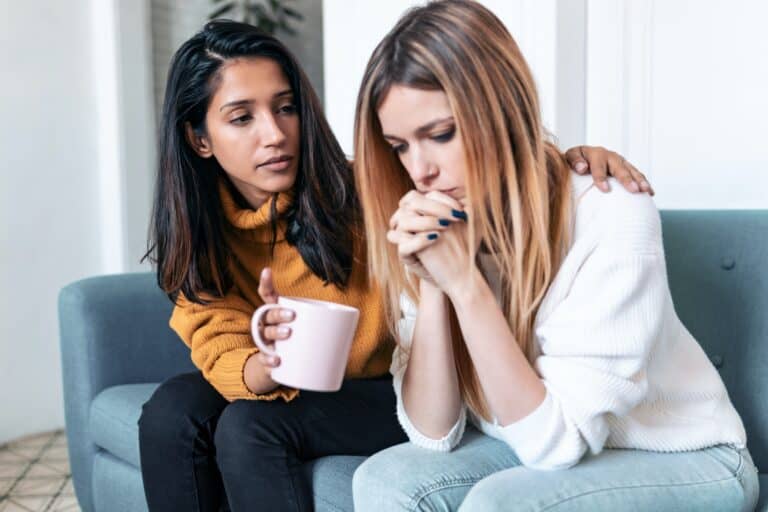 A woman with a hand on the shoulder of her worried friend as she comforts and supports her, an example of being there for someone who needs you