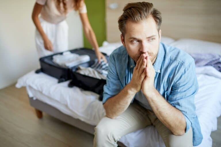 man sitting on bed in despair as wife packs suitcase to leave