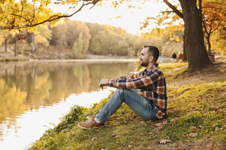 a happy guy with beard sitting alone on the grass in autumn park by the lake